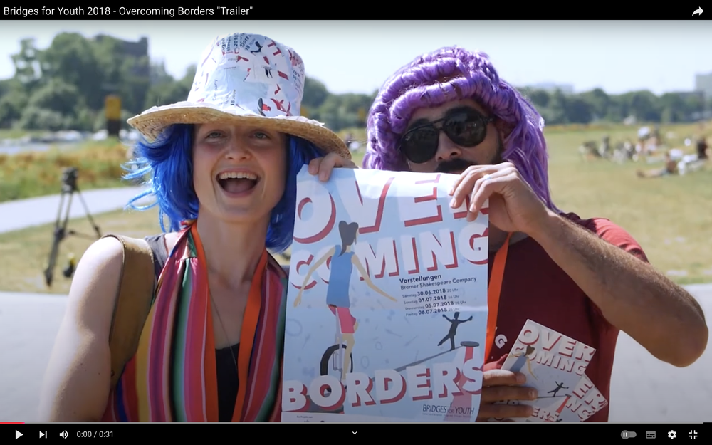 Screenshot von einem YouTube-Video. Zwei Personen stehen am Osterdeich in Bremen und halten das Plakat des Circusfestivals in die Kamera. Im Hintergrund ist verschwommen eine Wiese zu sehen, auf der Menschen liegen. Es ist Sommer und die Sonne scheint. Die Person links im Bild hat eine blaue Perücke auf dem Kopf und trägt einen Strohhut. Die Person rechts im Bild hat eine lila Perücke auf dem Kopf und trägt eine große dunkle Sonnenbrille. Das Plakat, das sie in die Kamera halten ist hellblau. Darauf zu sehen sind zwei illustrierte Personen. Eine davon balanciert mit einem Einrad auf einem Hochseil, die andere Person ist nur als Silhouette zu erkennen und balanciert ebenfalls auf dem Seil, aber ohne Rad. Der Text auf dem Plakat lautet: Overcoming borders. In kleinerer Schrift sind dort die Termine der Vorstellungen in der Bremer Shakespeare Company abgedruckt.  