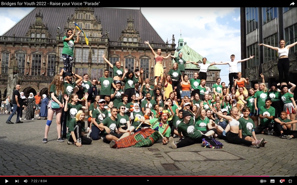 Screenshot von einem YouTube-Video. Das Bild zeigt die Gruppe der Teilnehmenden des Circusfestivals auf dem Bremer Marktplatz. Sie posieren für ein Gruppenfoto. Es sind mehr als 50 Personen, viele von ihnen haben das grüne T-Shirt mit dem Logo des Festivals an. Einige stehen auf den Schultern von anderen und recken dir Arme in die Höhe. Im Vordergrund liegt eine Person mit Clownskostüm auf dem Boden. Ebenfalls im Vordergrund machen drei Jugendliche einen Spagat. Im Hintergrund ist das Bremer Rathaus zu erkennen, rechts das Gebäude der Bürgerschaft. Das Bild wurde aufgenommen bei der Circusparade, die die Gruppe in der Innenstadt gezeigt hat. 